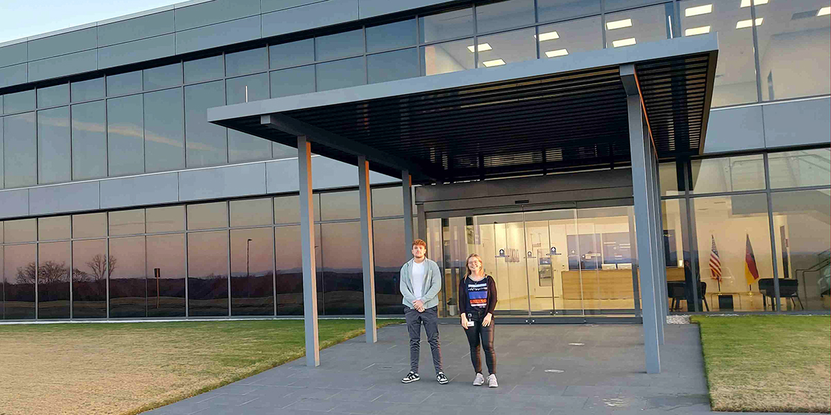 Gebäude von außen, davor zwei Mitarbeiter lachen in die Kamera