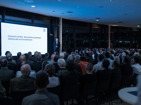 dunkler Saal mit vielen Menschen sitzen auf Stühlen ein Mann steht auf Bühne weiße Leinwand beleuchtet mit blauer Schrift "Globales Umfeld: Verunsicherung bremst Welthandel und Investitionen"