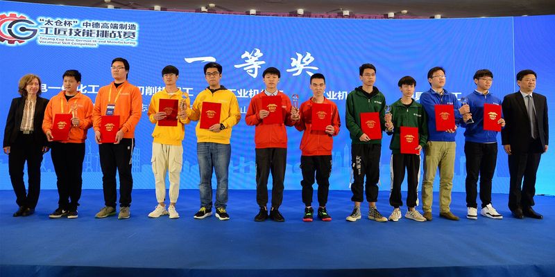 12 Personen asiatischer Herkunft stehen auf einer blauen Bühne, einige davon mit Urkunden und Pokalen in der Hand