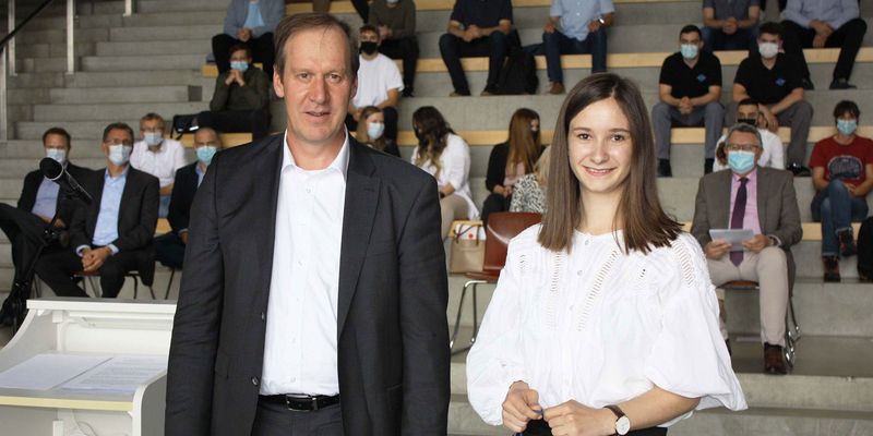 braunhaariger Mitarbeiter im Anzug mit junger Mitarbeiterin in einer weißen Bluse und hält eine Geschenktüte in der Hand