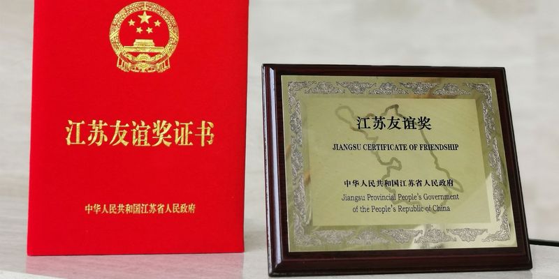 Freundschaftspreise, links rotes kleines Buch mit chinesischer goldener Aufschrift und rechts ein goldenes Bild mit holz Umrandung und schwarzer Schrift "Jiangsu Certificate of Friendship - Jiangsu Provincial People´s Government of the People´s Republic of China"
