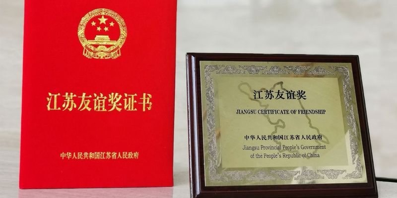 Freundschaftspreise links rotes kleines Buch mit chinesischer goldener Aufschrift rechts goldenes Bild mit Holt Umrandung schwarzer Schrift "Jiangsu Certificate of Friendship - Jiangsu Provincial People´s Government of the People´s Republic of China"