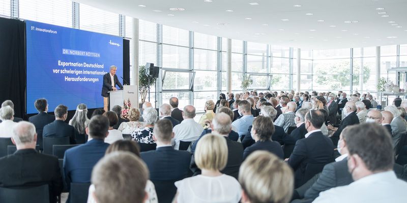 links Bühne mit männlichen Moderator blaue Leinwand weiße Schrift "Exportnation Deutschland vor schwierigen internationalen Herausforderungen" rechts Publikum 