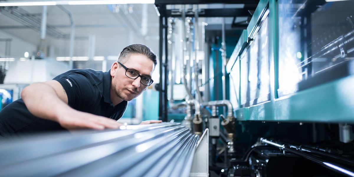 dunkelhaariger Mitarbeiter mit Brille an einer Maschine, kontrolliert Metallteile