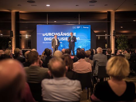 Vordergrund Publikum Hintergrund Geschäftsleitung Jürgen Häring mit dunkelhaarigem Mann auf Bühne dahinter blaue Leinwand mit weißer Aufschrift "Durchgängige Digitalisierung"