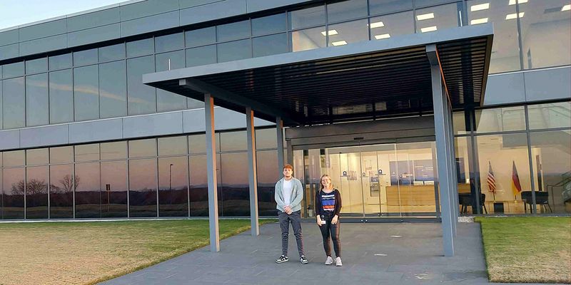 Gebäude von außen, davor zwei Mitarbeiter lachen in die Kamera