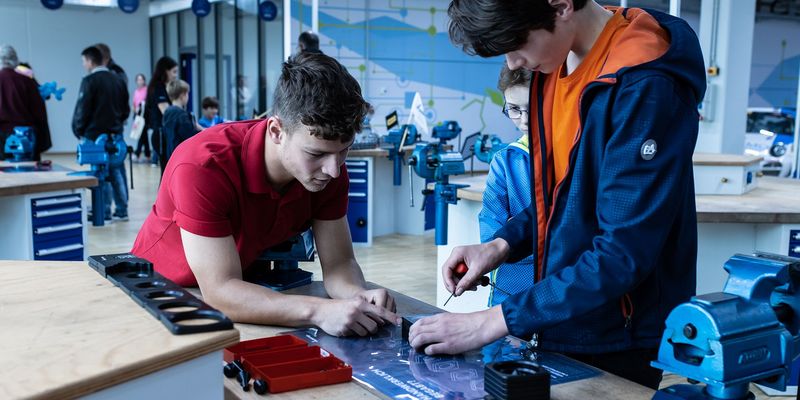 Links steht ein Azubi mit rotem T-Shirt angelehnt an einer Werkbank und zeigt zwei Schülern ein Metallteil