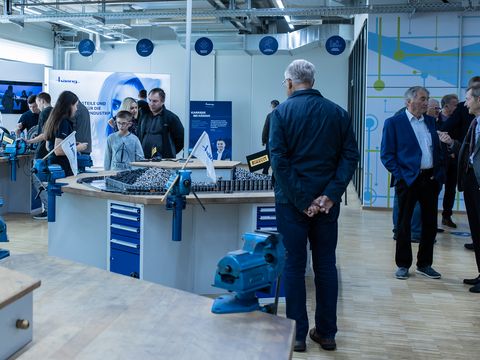 Eltern und Kinder stehen an Werkbänken und schauen sich um, Mitarbeiter/innen von Häring stehen bei den Kindern